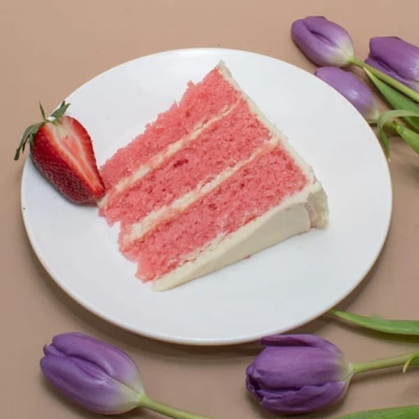 A slice of pink cake with white frosting and a strawberry on a white plate with purple tulips.