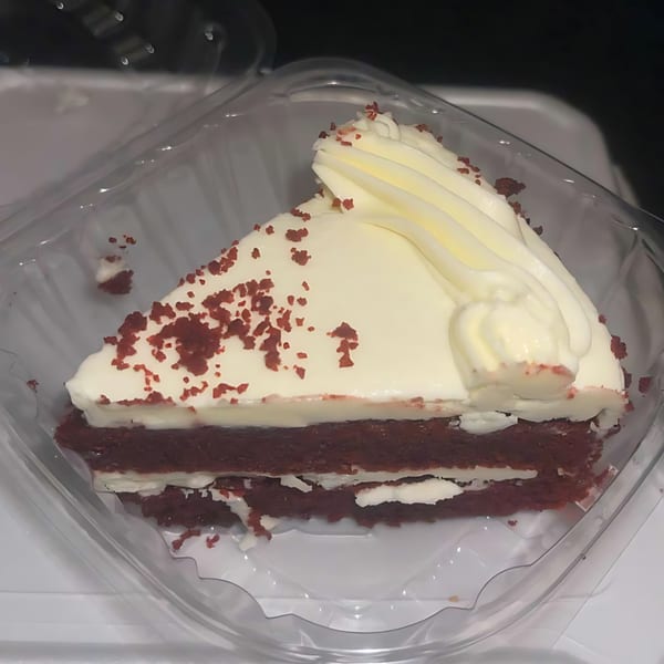 A slice of red velvet cake with white frosting and red sprinkles in a clear plastic container.