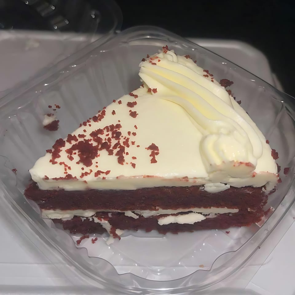 A slice of red velvet cake with white frosting and red sprinkles in a clear plastic container.