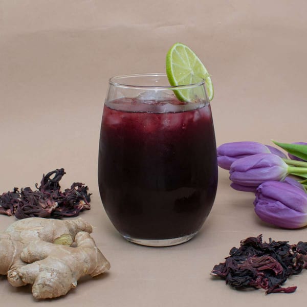 A glass of dark red Caribbean sorrel drink with ice and a lime wedge, surrounded by ginger and dried hibiscus.